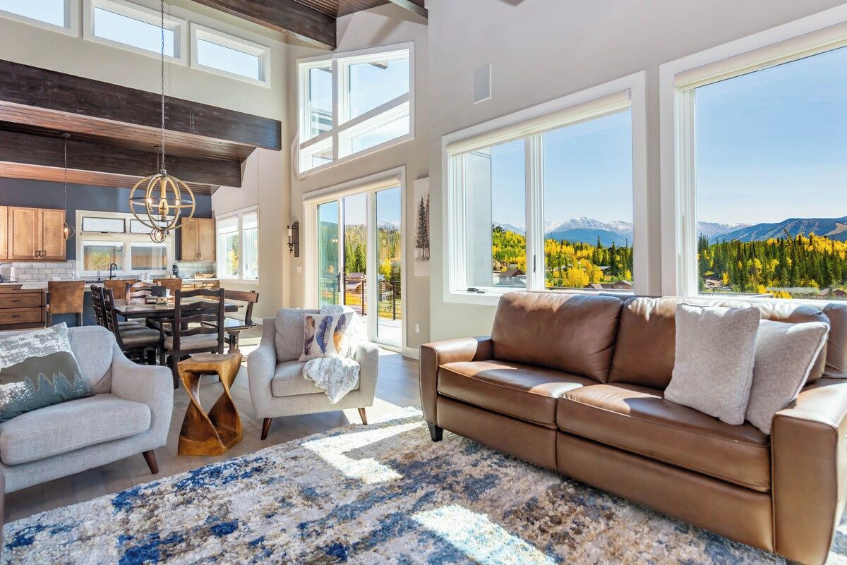 Un vaste espace de vie avec une grande fenêtre donnant sur un paysage montagneux, un coin salon confortable avec un canapé en cuir marron et une cuisine moderne avec des poutres en bois.