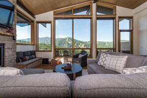 A spacious living room with a stone fireplace, large windows offering mountain views, and comfortable seating. - Lakota Apex | Close To Skiing! | Private Hot Tub w/ Resort Views (Winter Park)
