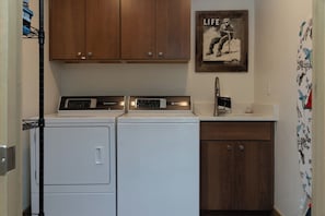 Ett tvättrum med två tvättmaskiner och torktumlare placerade ovanpå varandra, träskåp ovanför och en affisch med ett livstema på väggen.