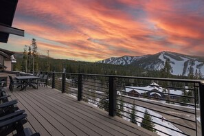 Une terrasse en bois avec des sièges d’extérieur, offrant une vue sur un paysage montagneux aux sommets enneigés et sur un ciel au coucher du soleil.
