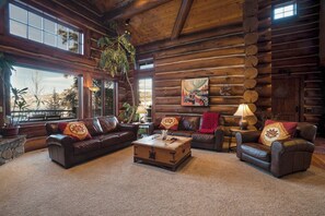 A log cabin interior with a fireplace, leather sofas, a wooden coffee table, and large windows.