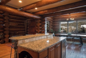 A log cabin interior with a kitchen island, wooden beams, and a view of the outdoors. - Moose Drool Lodge | Luxury Mountain Lodge | Private Hot Tub (Winter Park)