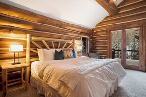 A log cabin bedroom with a large bed, wooden headboard, and a view of a snowy forest through a sliding glass door.