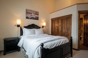 Quarto com cama, duas luminárias de cabeceira, porta de madeira e um quadro na parede.