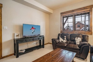 Sala de estar com televisão, sofá de couro, mesa de centro de madeira e janela com vista para uma casa.