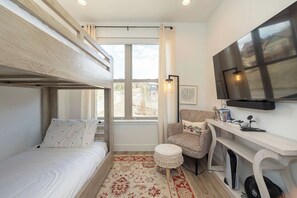 Ein Zimmer mit einem Etagenbett, einem Schreibtisch mit Computer, einem Sessel, einem Fernseher und einem Fenster mit Blick auf Gebäude.