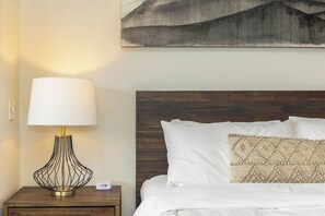 Ein ordentlich bezogenes Bett mit weißer Bettwäsche und einem dekorativen Kissen, eine Nachttischlampe mit einem einzigartigen Drahtgestelldesign und ein Wandbild über dem Bett.