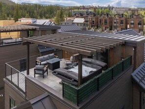 A rooftop terrace with a hot tub, seating area, and a view of surrounding buildings. - Gather & Glow | Downtowan House | Private Hot Tub (Winter Park)