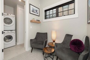 A laundry room with a washing machine, two grey armchairs, a small wooden table with a lamp, and a window with a view of a building. - Gather & Glow | Downtowan House | Private Hot Tub (Winter Park)