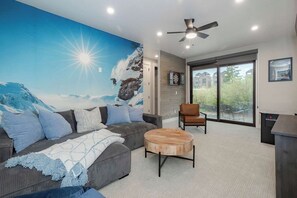 A modern living room with a large mural, a ceiling fan, and a view through glass doors. - Gather & Glow | Downtowan House | Private Hot Tub (Winter Park)