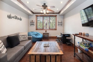 Una sala de estar con sofá, mesa de centro, sillón de pie, televisor y ventana con vistas a árboles.