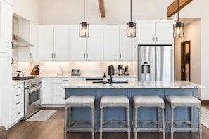 A modern kitchen with white cabinetry, a stainless steel refrigerator, and a marble island with bar stools. - Lakota Evergreen | Slope Views | Private Hot Tub (Winter Park)