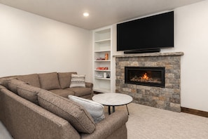 A living room with a stone fireplace, a flat-screen TV mounted above it, a sectional sofa, and a small round coffee table. - Lakota Evergreen | Slope Views | Private Hot Tub (Winter Park)