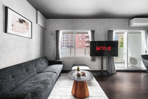 A modern living room with a grey sofa, a coffee table with decorative items, a flat-screen TV displaying 'NETFLIX', and a view of a red building through the window. - Edgewater Terrace 211 (Sapporo)