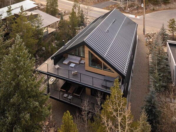 A modern two-story house with a sloped roof, a covered deck, and a large window. - Creekside Retreat | Private Hot Tub | Secluded Modern Retreat (Winter Park)