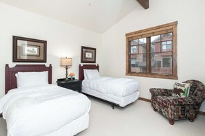 Ein Schlafzimmer mit zwei Betten, einem Nachttisch mit Lampe, einem Sessel mit einem Kissen mit der Aufschrift 'Lodge' und einem Fenster mit Blick auf Gebäude.
