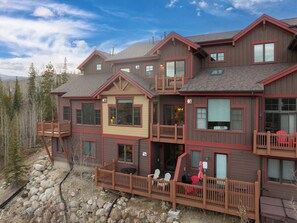 A multi-story building with balconies, a stone retaining wall, and a wooden deck with chairs. - Bear Den | Private Deck | Rocky Mountain Views (Winter Park)