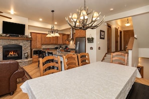 A spacious living area with a fireplace, a dining table with chairs, a kitchen with wooden cabinets, and a staircase. - Bear Den | Private Deck | Rocky Mountain Views (Winter Park)