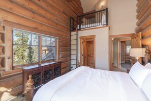 Ein Schlafzimmer im Blockhausstil mit Schlafplatz in der Mansarde, einem Fenster mit Blick auf Bäume und einer Holzkiste.