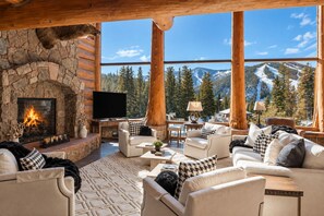 A log cabin interior with a stone fireplace, a flat-screen TV, and a view of snow-capped mountains. - Lakota Cedarcrest | Large & Luxe | Private Hot Tub | Resort Views (Winter Park)