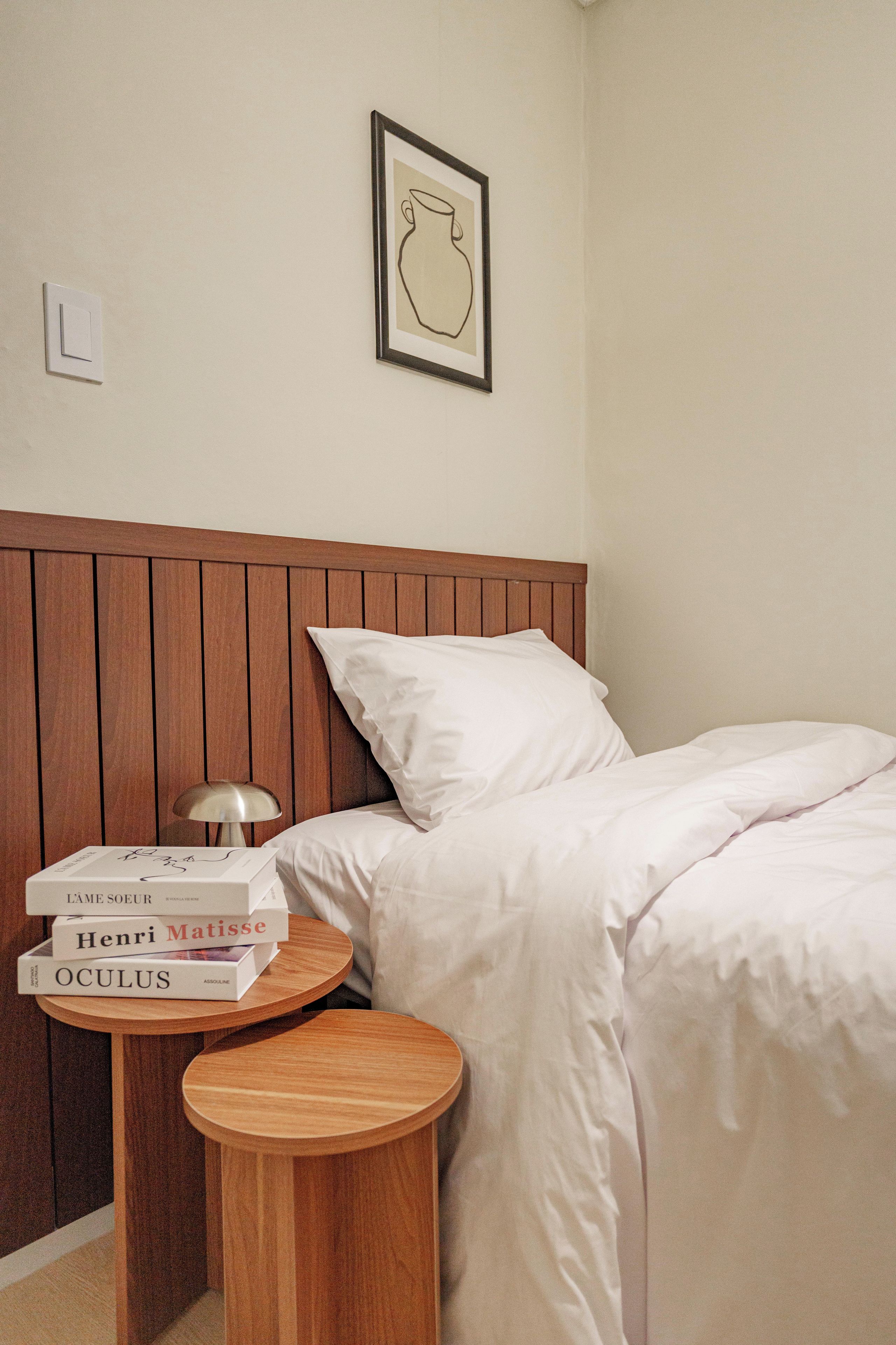 Una cama bien hecha con sábanas blancas, un cabecero de madera y una mesita de noche con una lámpara y libros.