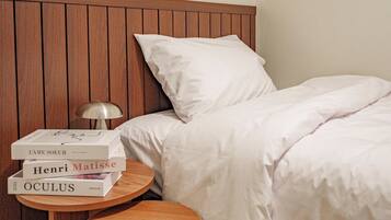 Una cama bien hecha con sábanas blancas, un cabecero de madera y una mesita de noche con una lámpara y libros.