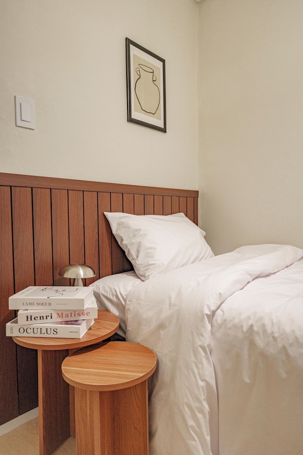 Una cama bien hecha con sábanas blancas, un cabecero de madera y una mesita de noche con una lámpara y libros.