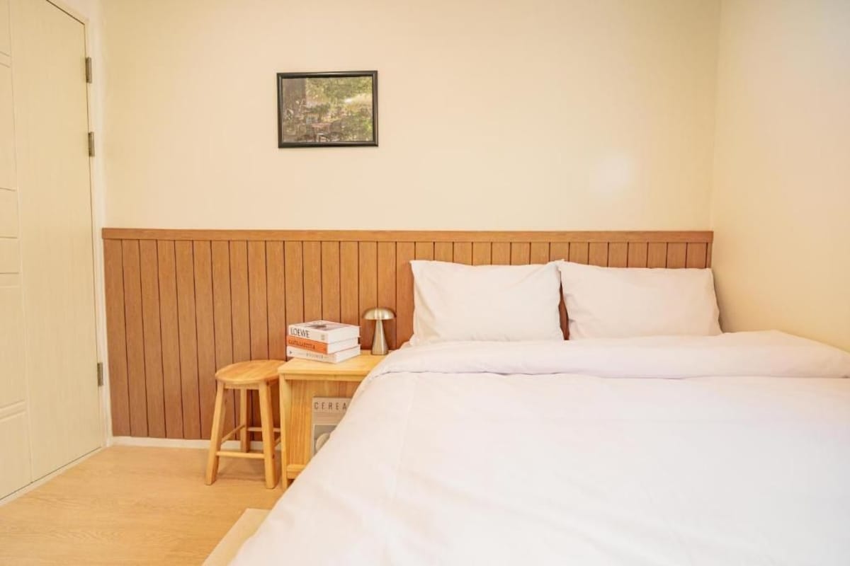 Una camera da letto con una testiera in legno, un letto con biancheria da letto bianca, un comodino con libri e una lampada, e un quadro appeso al muro.