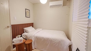 A small, neatly arranged bedroom with a single bed, a bedside table with books, a wall-mounted air conditioner, and a window with blinds. - WAYHOME (Seoul)