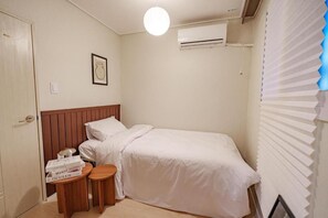 Una camera da letto piccola e ordinata con un letto singolo, un comodino con dei libri, un condizionatore d'aria a muro e una finestra con persiane.