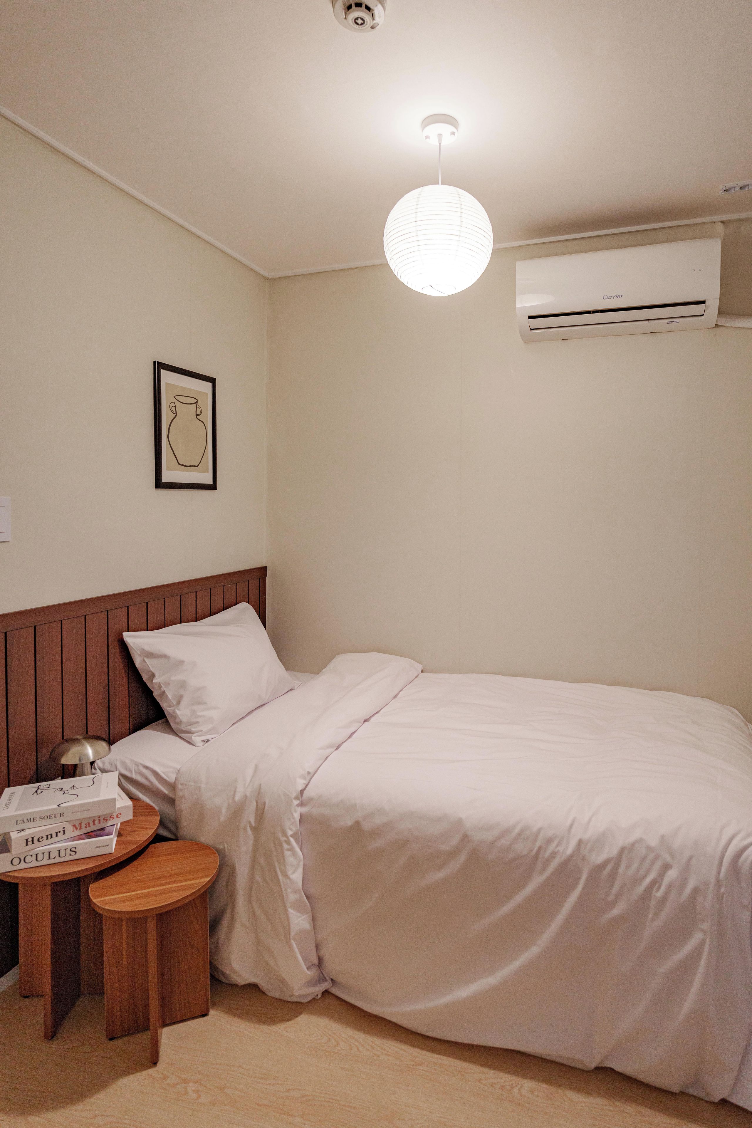 A small, neatly made room with a single bed, a bedside table with books, a wall-mounted air conditioner, and a simple light fixture.