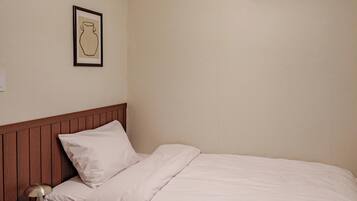 A small, neatly made room with a single bed, a bedside table with books, a wall-mounted air conditioner, and a simple light fixture.