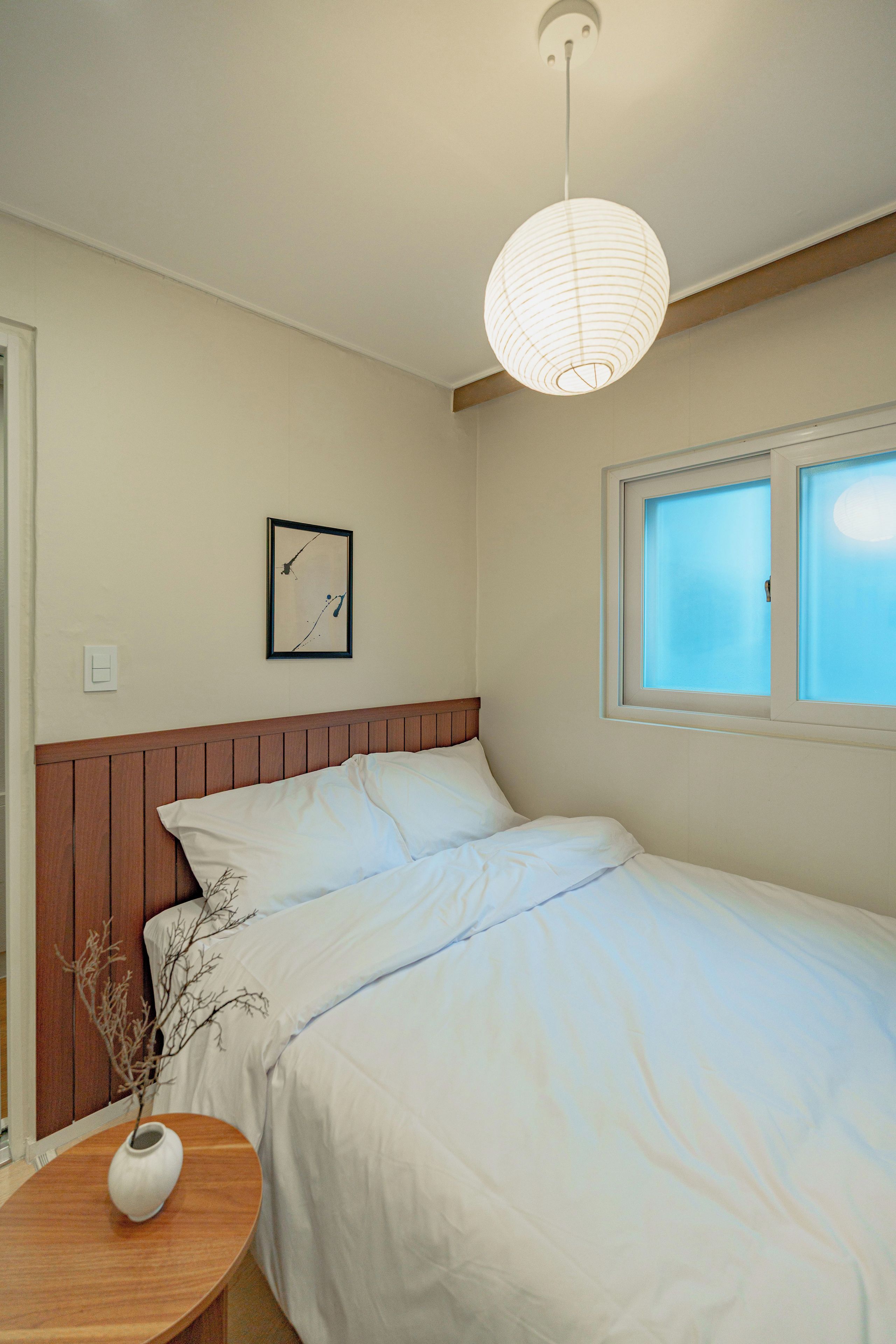 A bedroom with a single bed, a bedside table with a vase, a framed picture on the wall, and a window with blinds.