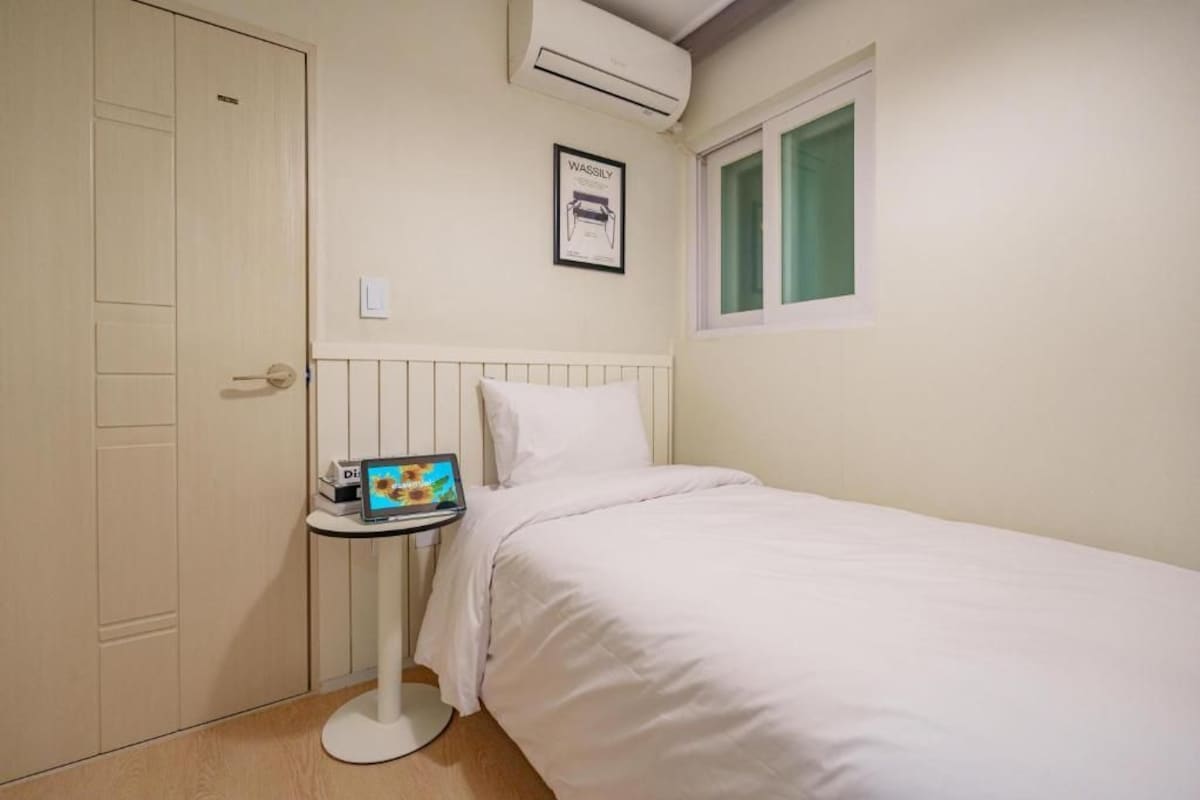 Una camera da letto con un letto, un comodino con un portatile, una finestra con tende e un condizionatore d'aria.