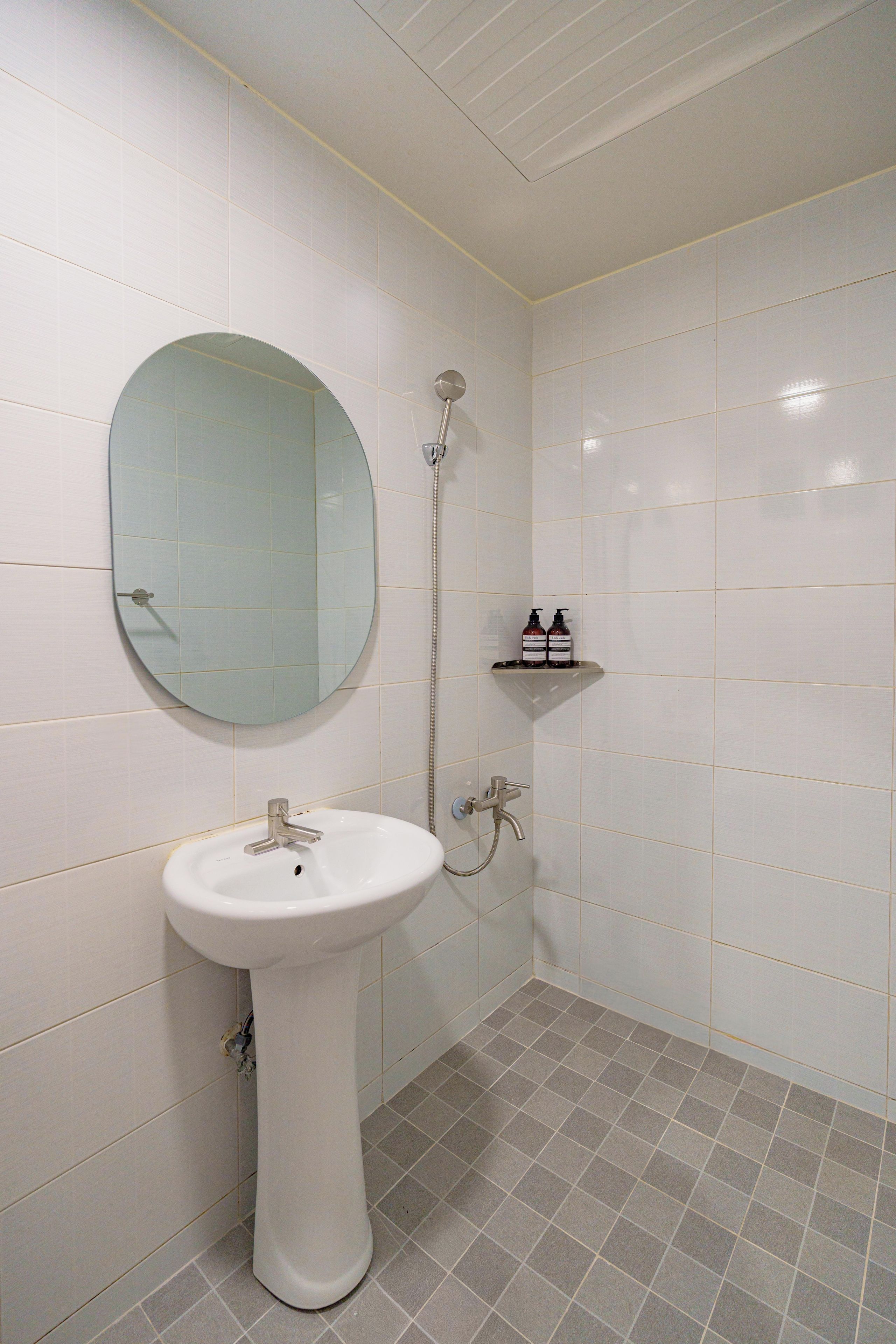 Un baño con un lavamanos blanco, un espejo redondo, una ducha y un estante con artículos de aseo.