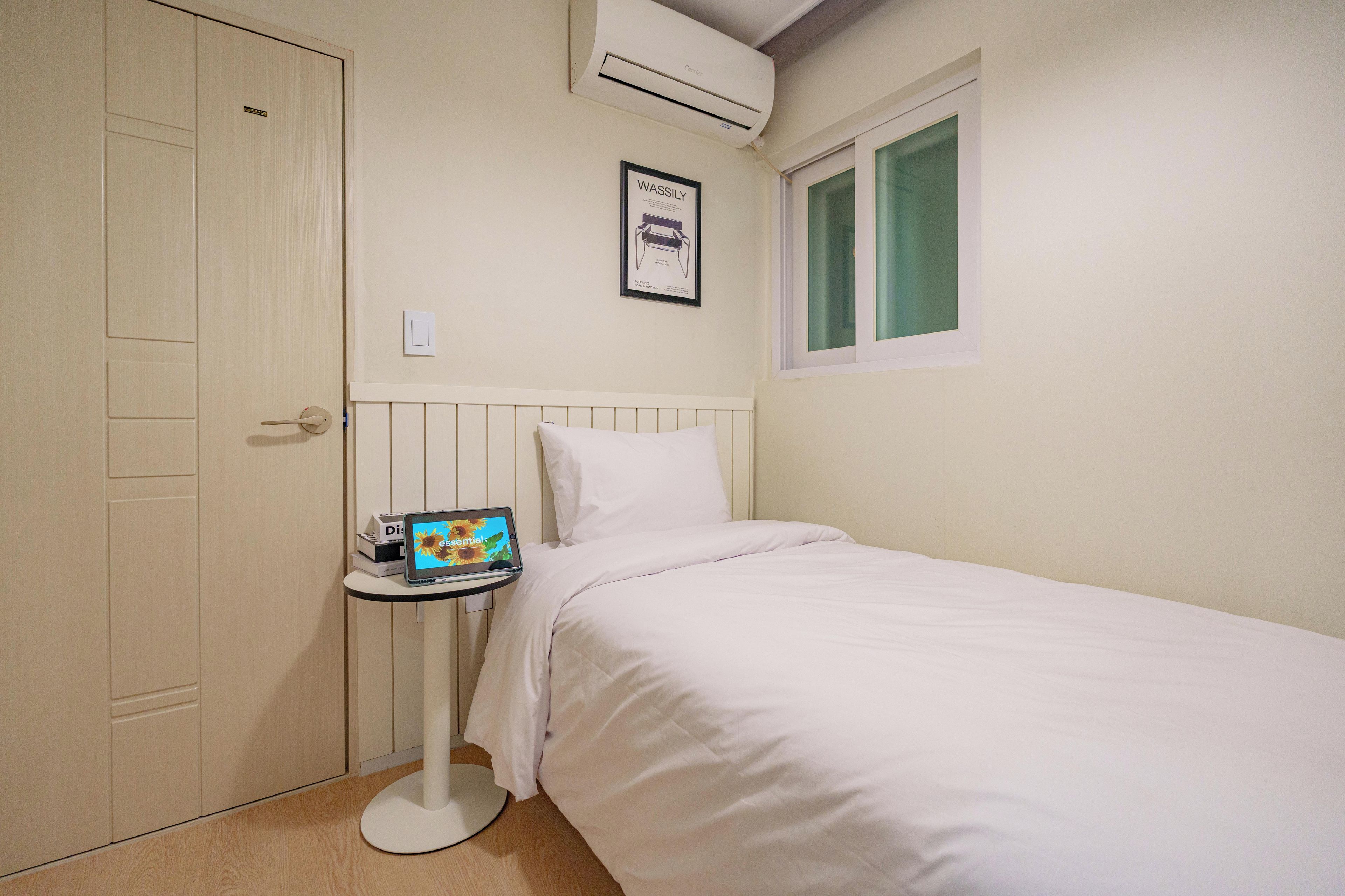 Un dormitorio con cama, una mesita de noche con una computadora portátil, una ventana y un equipo de aire acondicionado.