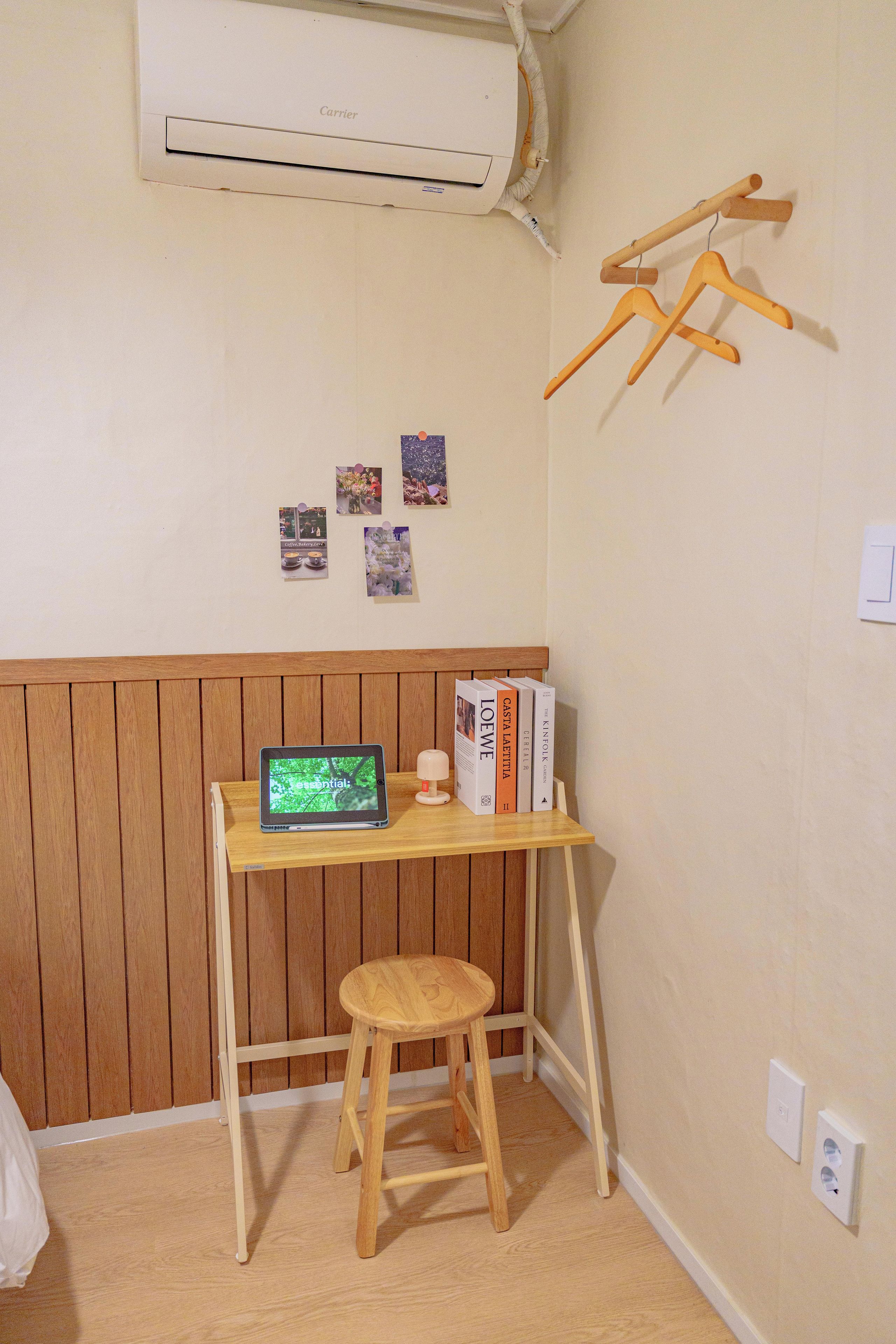 Una habitación pequeña con un escritorio de madera, un taburete, un aire acondicionado de pared y algunas fotografías.