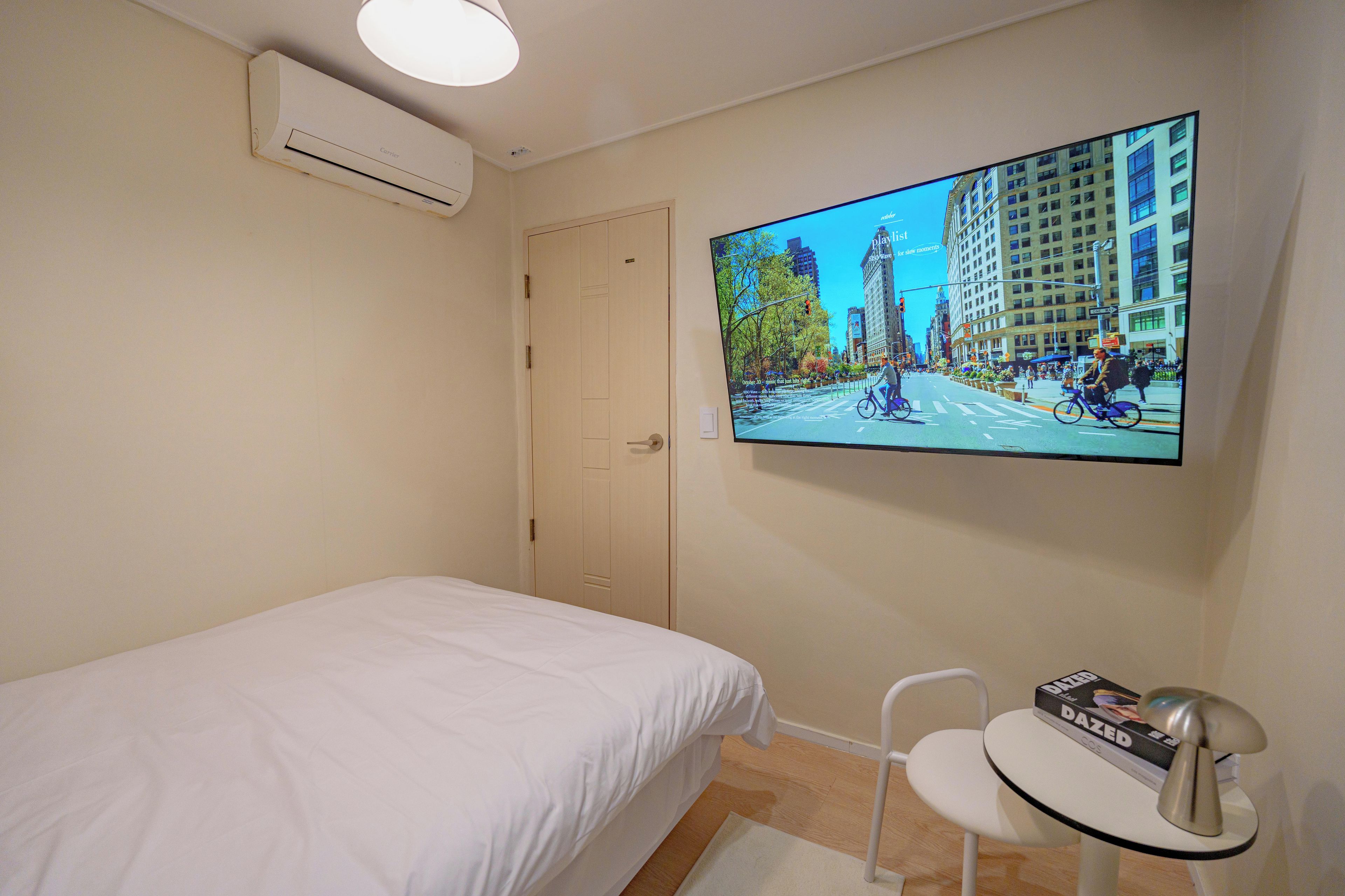 A hotel room with a bed, a desk, a chair, a flat-screen TV mounted on the wall, and an air conditioning unit.