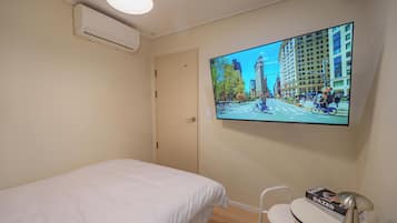 A hotel room with a bed, a desk, a chair, a flat-screen TV mounted on the wall, and an air conditioning unit.