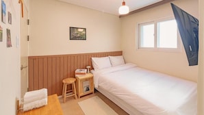 A single-bed room with a wooden headboard, a bedside table with a lamp, a small stool, and a window with a curtain. - WAYHOME (Seoul)