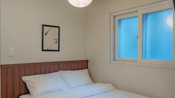 A bedroom with a bed, a wooden headboard, a window with blinds, a wall clock, and a hanging light fixture. - WAYHOME (Seoul)