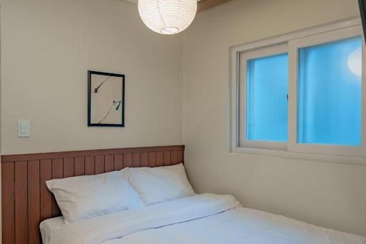 Una camera da letto con un letto, una testiera in legno, una finestra con persiane, una sveglia a parete e una lampada a sospensione.
