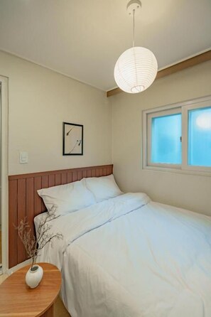 A bedroom with a bed, a bedside table with a vase, a wall clock, and a window with blinds. - WAYHOME (Seoul)