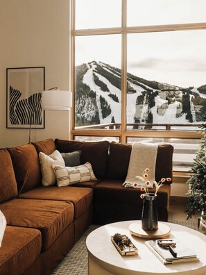 Ein Wohnzimmer mit einem braunen Ledersofa, einem runden Couchtisch mit einer Vase getrockneter Blumen und einem großen Fenster mit Blick auf einen verschneiten Berg.