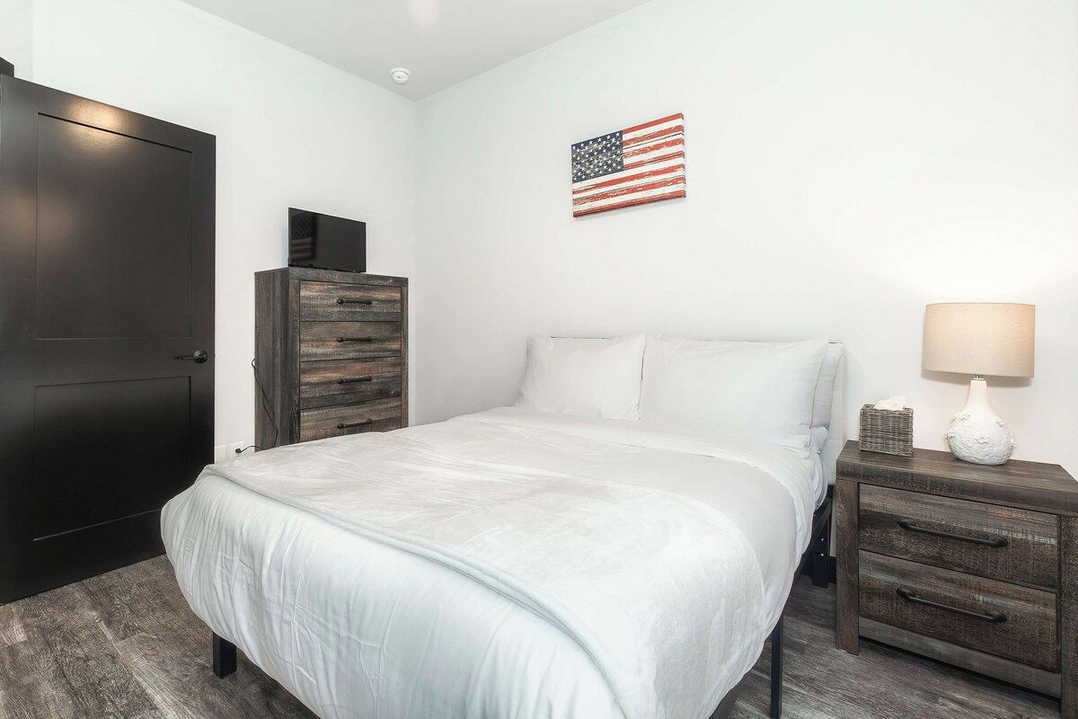 A bedroom with a bed, a nightstand, a dresser, a wall-mounted TV, and an American flag.