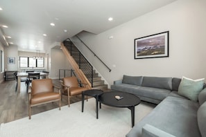 Un salon moderne avec un canapé gris, des fauteuils en cuir, une table basse et un escalier menant à un niveau supérieur.