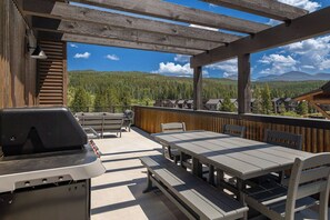An outdoor patio with a barbecue grill, wooden tables, and benches, overlooking a forested mountain area. - Peaks and Pines Villa | Downtown | Private Hot Tub (Winter Park)