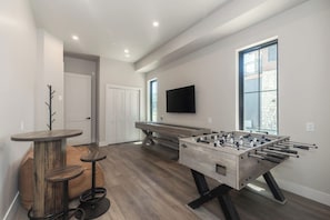 A room with a foosball table, a bar area with stools, a television mounted on the wall, and a door leading to another room. - Peaks and Pines Villa | Downtown | Private Hot Tub (Winter Park)