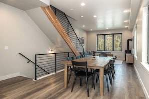 A dining area with a wooden table and chairs, a staircase with a railing, and a window with a view of trees. - Peaks and Pines Villa | Downtown | Private Hot Tub (Winter Park)