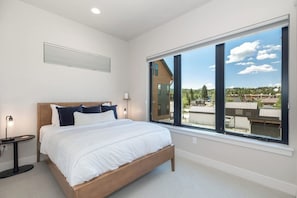 A modern bedroom with a large window, a bed, a bedside table, and a wall-mounted light fixture. - Peaks and Pines Villa | Downtown | Private Hot Tub (Winter Park)